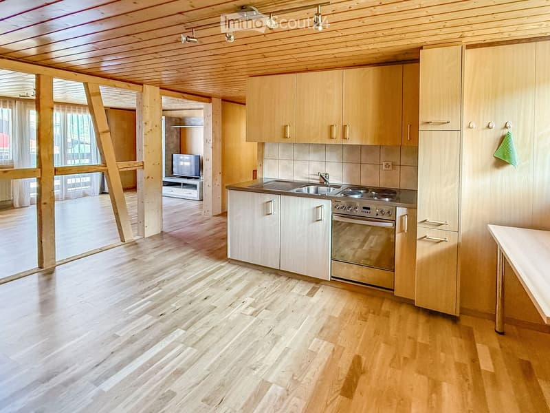 Modern kitchen with light wooden cabinets, stainless steel appliances, and a large island. The space features a wooden floor and ample natural light from large windows.