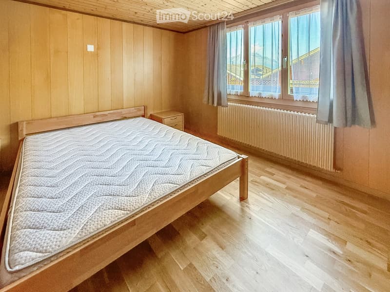 Cozy bedroom with wooden walls and ceiling, wooden floor, large windows with curtains, radiator, and a neatly made bed with a nightstand.
