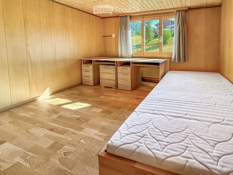Cozy bedroom with wooden walls and floor, featuring a single bed, a wooden desk with drawers, and a radiator beneath a large window with curtains.