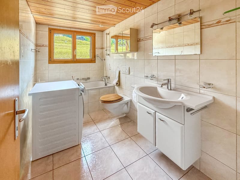 Modern bathroom with a bathtub, toilet, sink, and a large mirror above the sink. The room features tiled walls and a wooden ceiling.