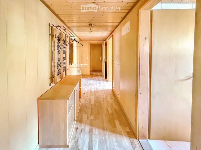 Bright hallway with wooden flooring, light-colored walls, and a coat rack with hangers on the left side. The hallway features several doors leading to other rooms and a small bench or table near the coat rack.