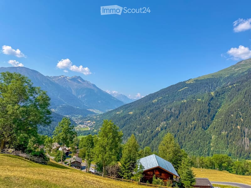 A picturesque mountain valley with scattered wooden houses, lush green trees, and a winding road, surrounded by towering peaks under a clear blue sky.