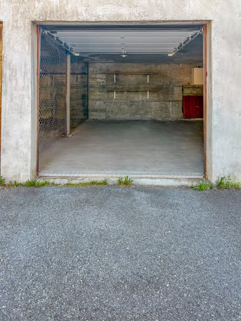 Open garage with concrete walls, metal door, and small storage area with shelves and boxes.