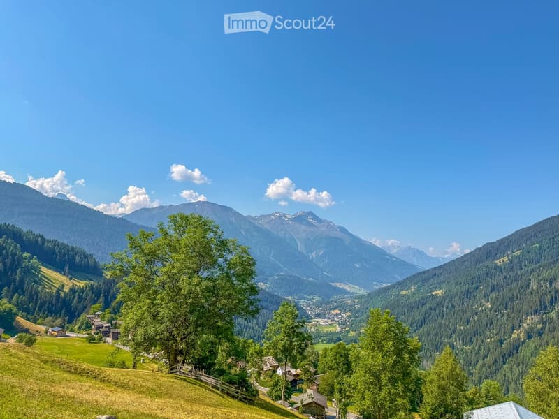 A panoramic view of a lush green valley surrounded by towering mountains, with scattered houses and trees dotting the landscape.
