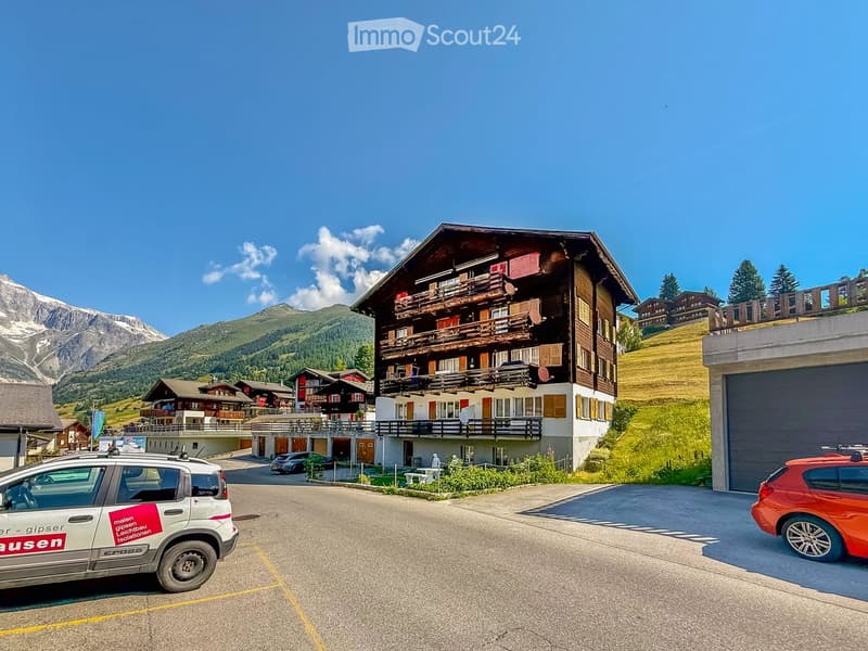 Multi-story wooden building with balconies and red shutters, surrounded by a parking area with cars and a scenic mountain backdrop.