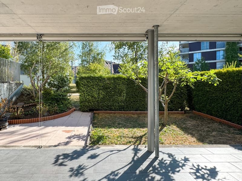 A modern outdoor patio area with a tiled floor, a covered pergola, a pathway leading to a garden, and surrounding greenery including trees and shrubs.
