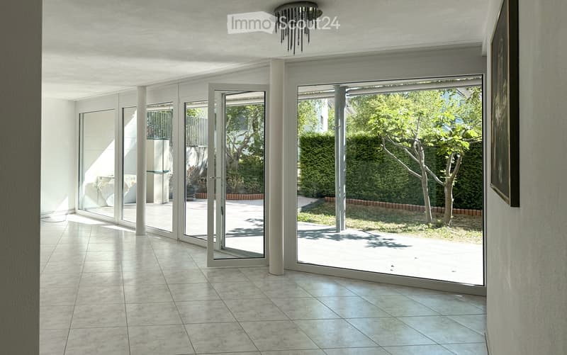 Bright, minimalist hallway with large glass sliding doors leading to a garden, white walls, tiled floor, and a modern chandelier.