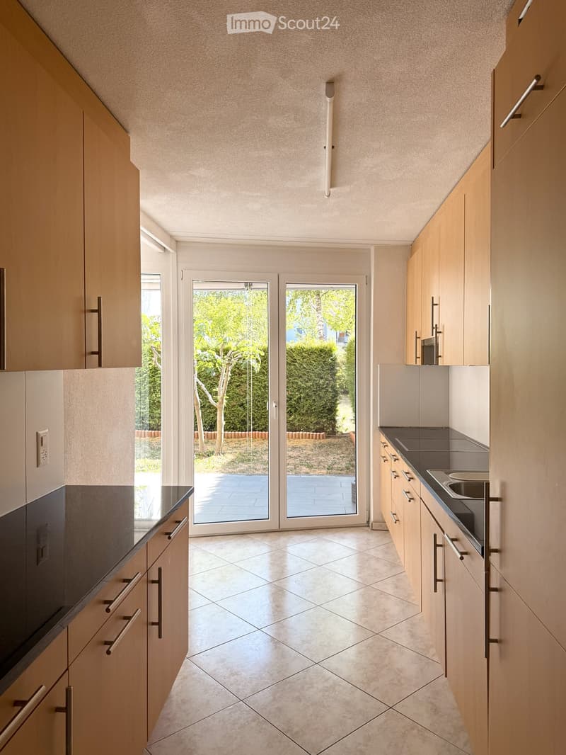 Modern kitchen with light wooden cabinets, dark countertops, and a large sliding glass door leading to a garden.