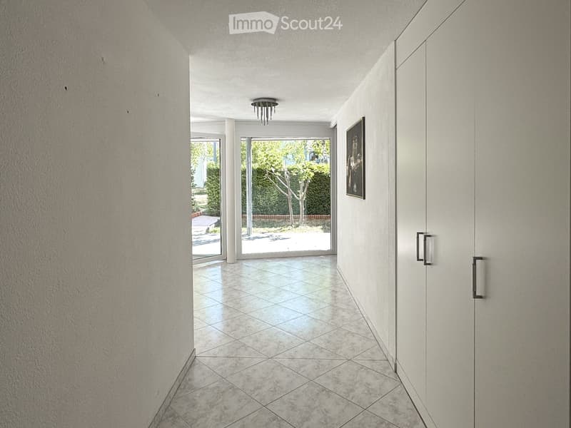 Modern hallway with tiled floor, white walls, recessed lighting, and a framed picture on the wall. The hallway leads to a bright outdoor area with greenery visible through large glass doors.