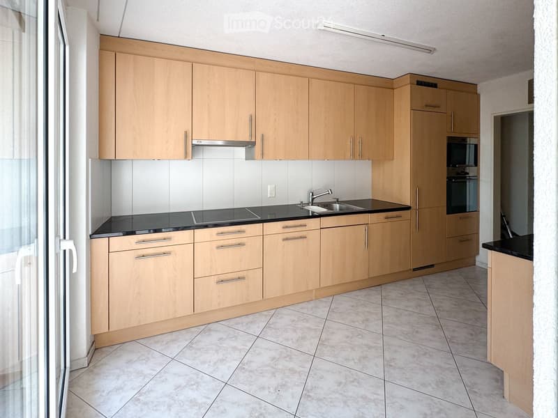 Modern kitchen with light wooden cabinets, black countertops, and stainless steel appliances including a sink and oven.