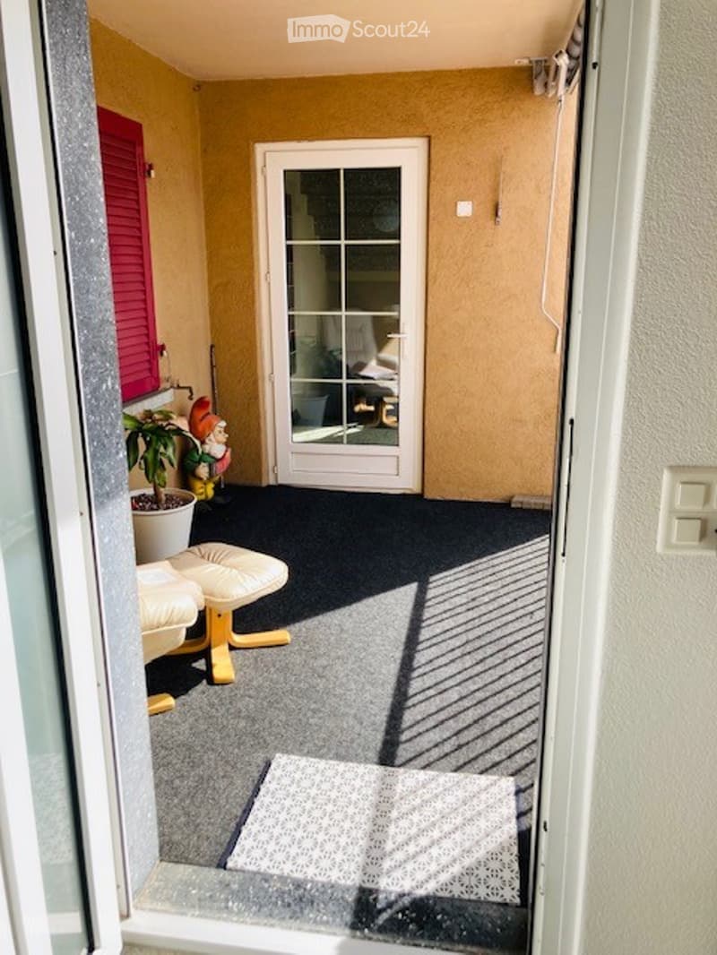 Balcony with grey carpet, white door leading inside, small potted plant, and two white cushioned stools.