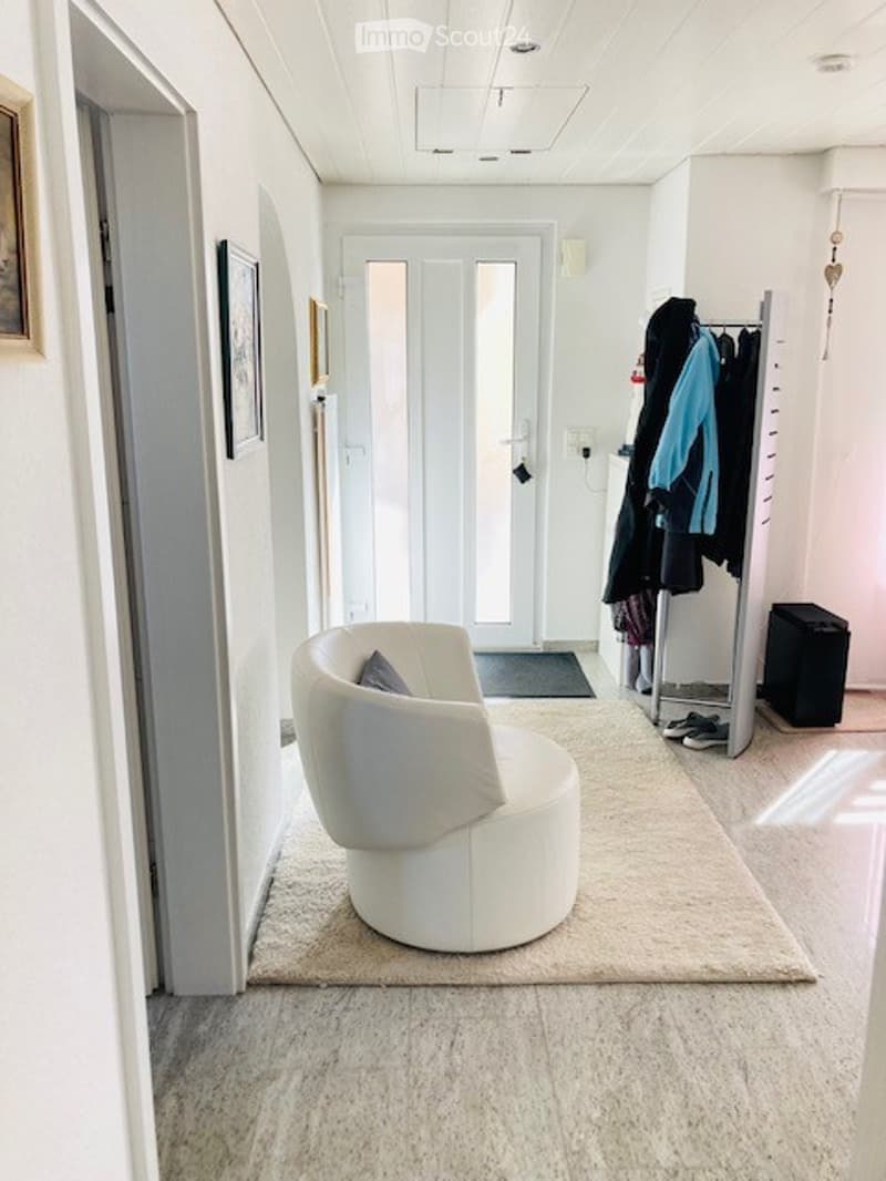 A modern, minimalist hallway with a white leather chair, a coat rack with jackets, and a small rug. The space features light-colored walls, a white door, and a small mirror on the wall.