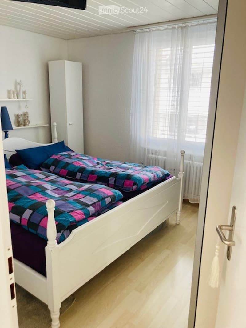 Cozy bedroom with a white wooden bed, colorful checkered bedspread, matching pillows, a white wardrobe, a small shelf with decorative items, and a blue lamp on a nightstand.