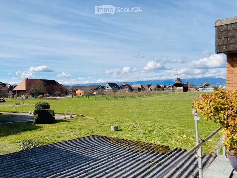 A peaceful rural landscape with green fields, scattered houses, and distant mountains under a clear blue sky.