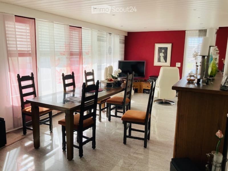 Bright dining area with a glass dining table, six wooden chairs, a television, a white sofa, and decorative items including a flower vase and picture frame.