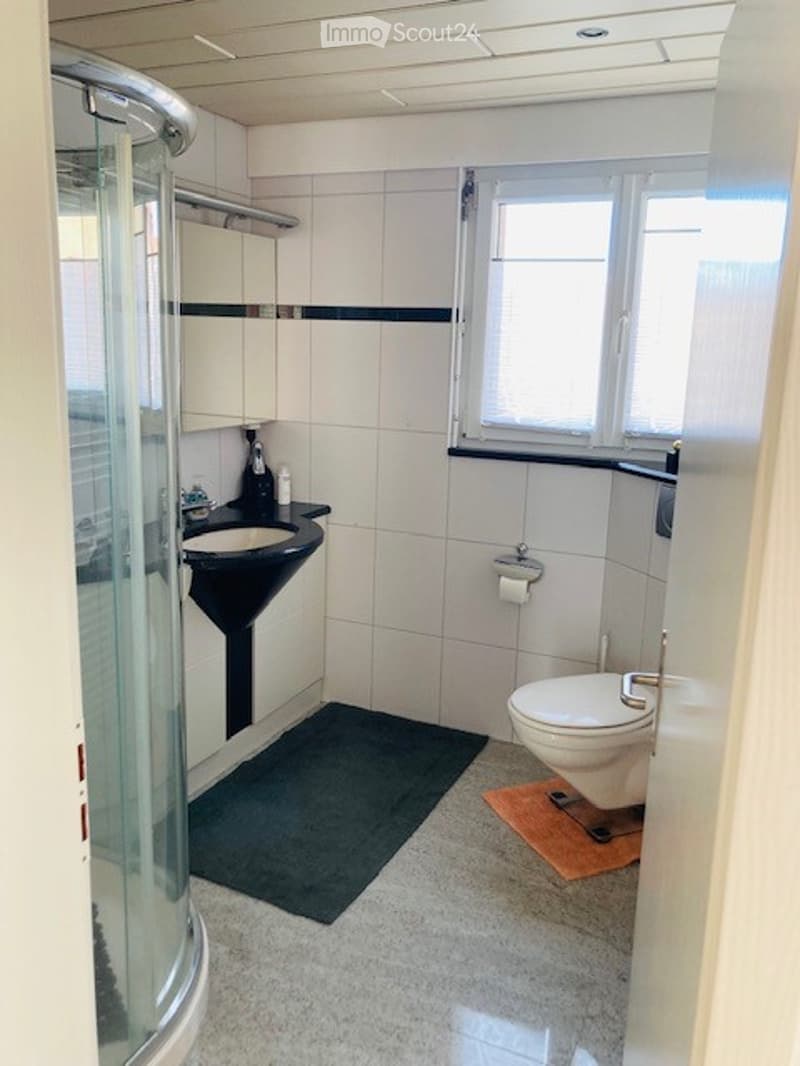 Modern bathroom with white tiles, black sink, toilet, shower, and a small window for natural light.