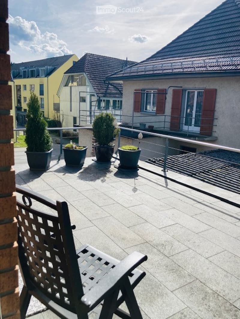Balcony with tiled floor, metal railing, potted plants, and a single wooden chair.