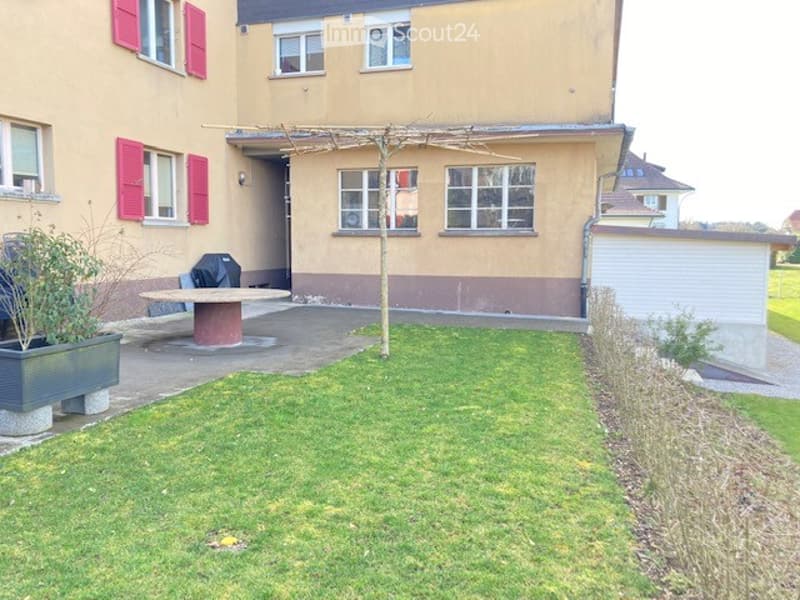 Well-maintained garden with a small patio area, a table, a bench, and a planter near the house. The house has red shutters and a visible garage.
