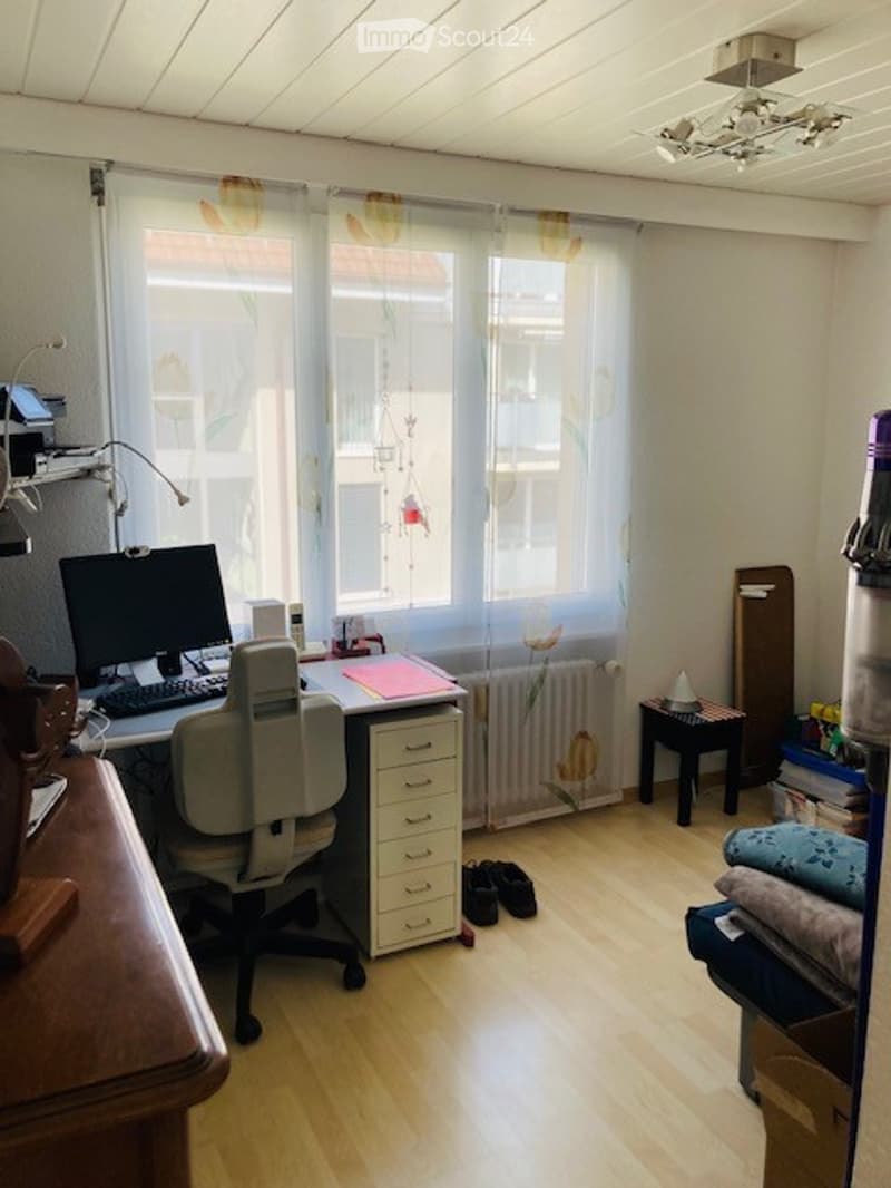 A furnished room with a desk, chair, computer, and cabinet. The room has wooden flooring, large windows with curtains, and a radiator under the window.