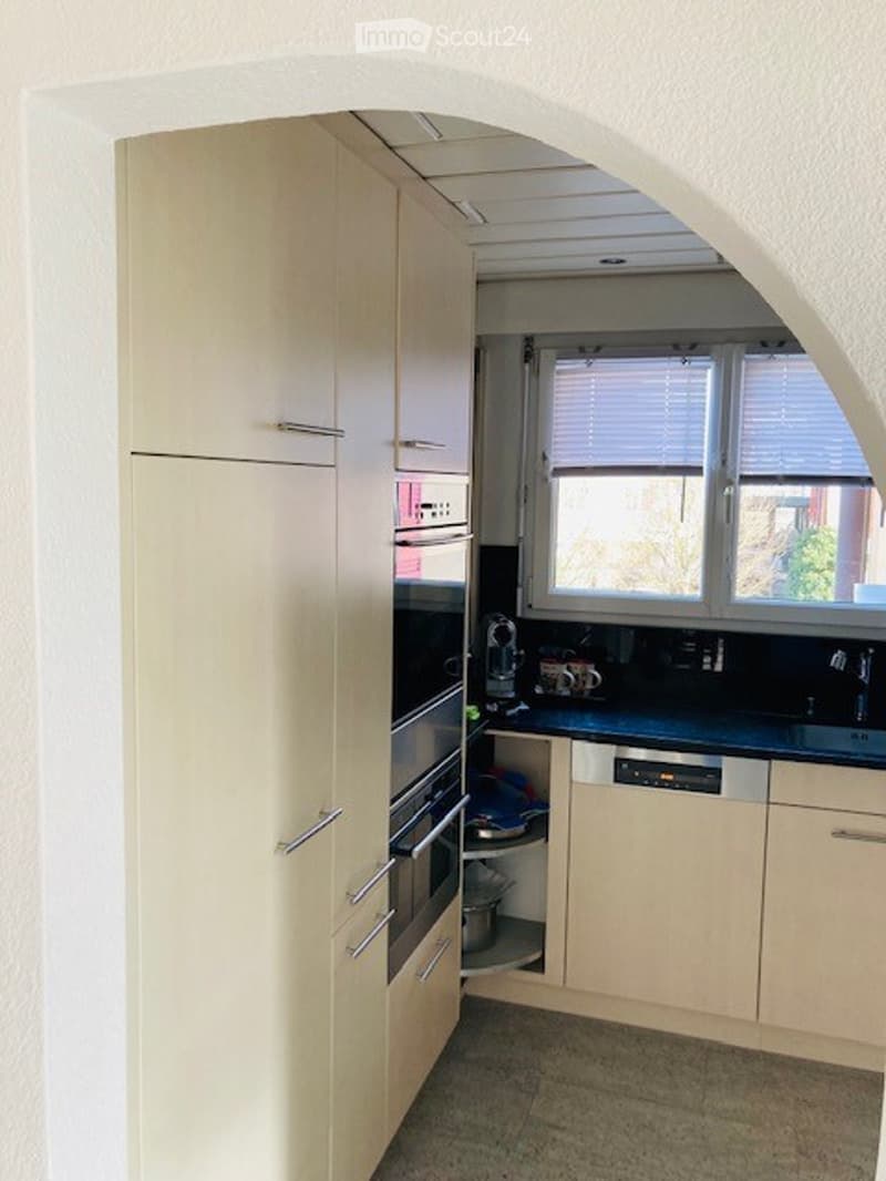 Modern kitchen with light-colored cabinets, stainless steel oven, dishwasher, and sink. Windows with blinds provide natural light.