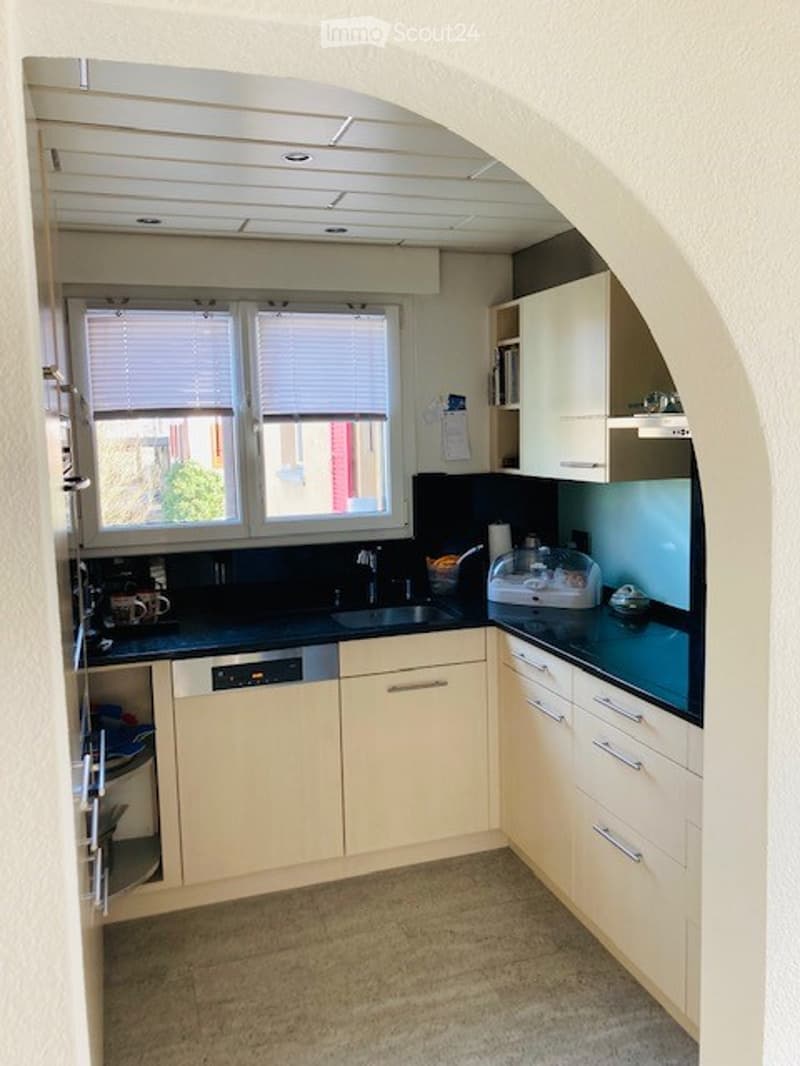 Modern kitchen with light-colored cabinets, black countertops, and stainless steel appliances. Features a sink, dishwasher, microwave, and refrigerator, with large windows and blinds.