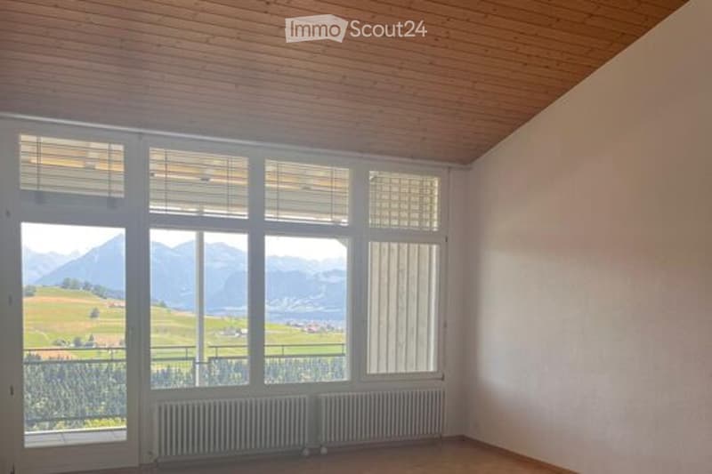 Empty room with large windows offering a scenic mountain view, wooden ceiling, and radiators.