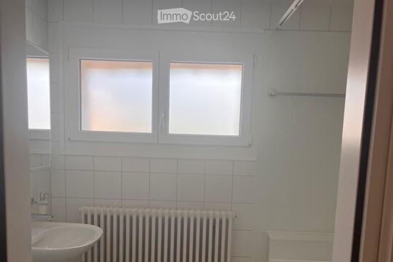 Bathroom with white tiled walls, a sink, a radiator, and a bathtub. Three windows with frosted glass panes provide natural light.