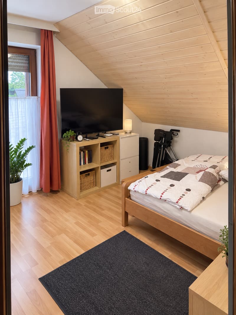 Cozy bedroom with wooden floors, slanted wooden ceiling, bed with patterned quilt, nightstand with lamp, bookshelf with books and baskets, potted plant, television, and a tripod in the corner.