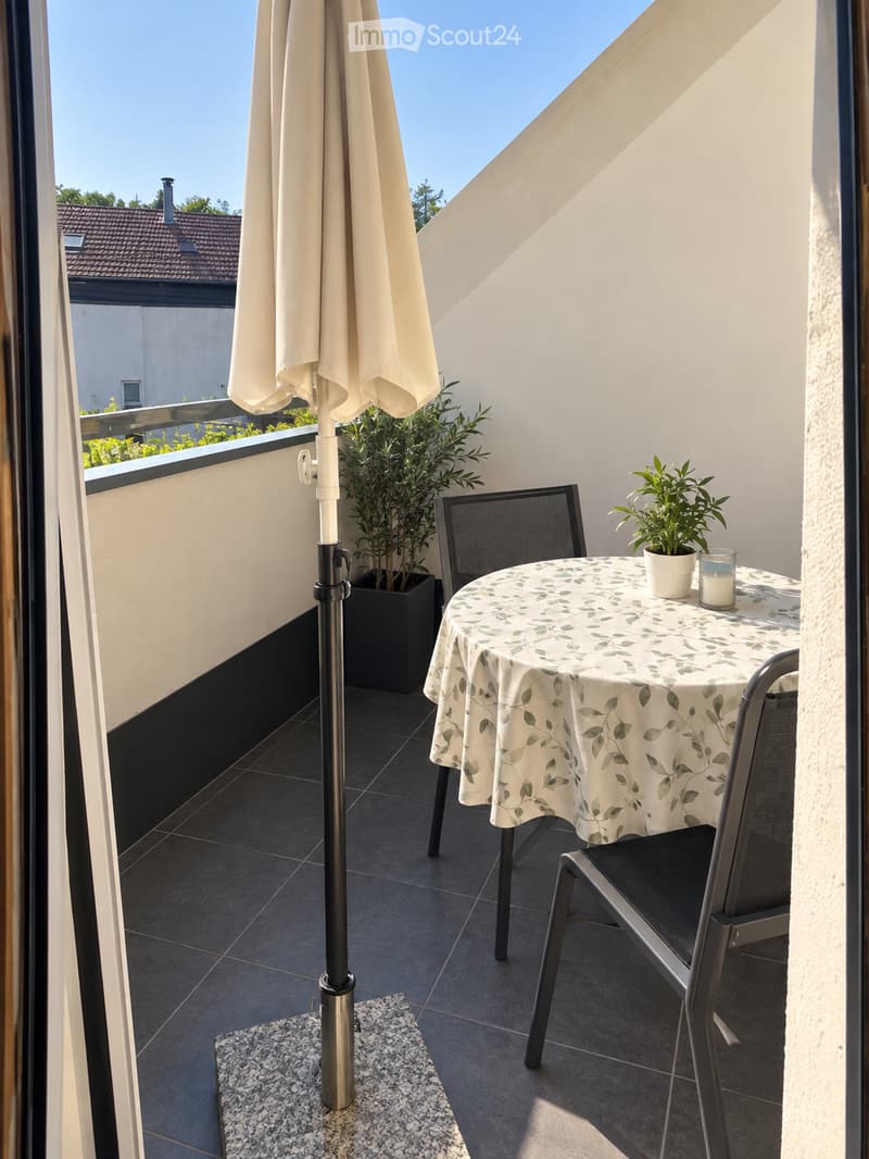 Balcony with a round table covered in a leaf-patterned cloth, two black metal chairs, a large umbrella pole, potted plants, and a candle on the table.