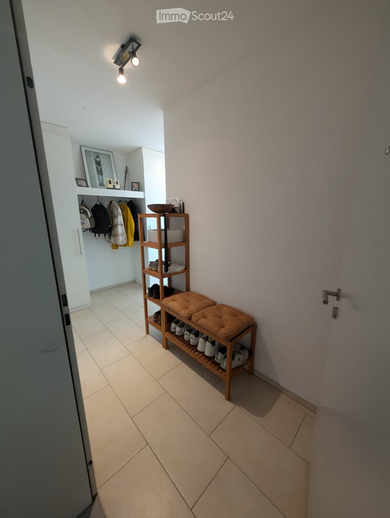 A well-lit hallway with tiled flooring, a wooden shoe rack, and a small bench. The hallway features built-in storage with coats and bags, along with framed artwork on the wall.