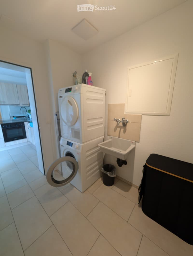 Compact bathroom with a stacked washing machine and dryer, a small sink, and a trash bin. The space is clean and well-lit with tiled flooring.