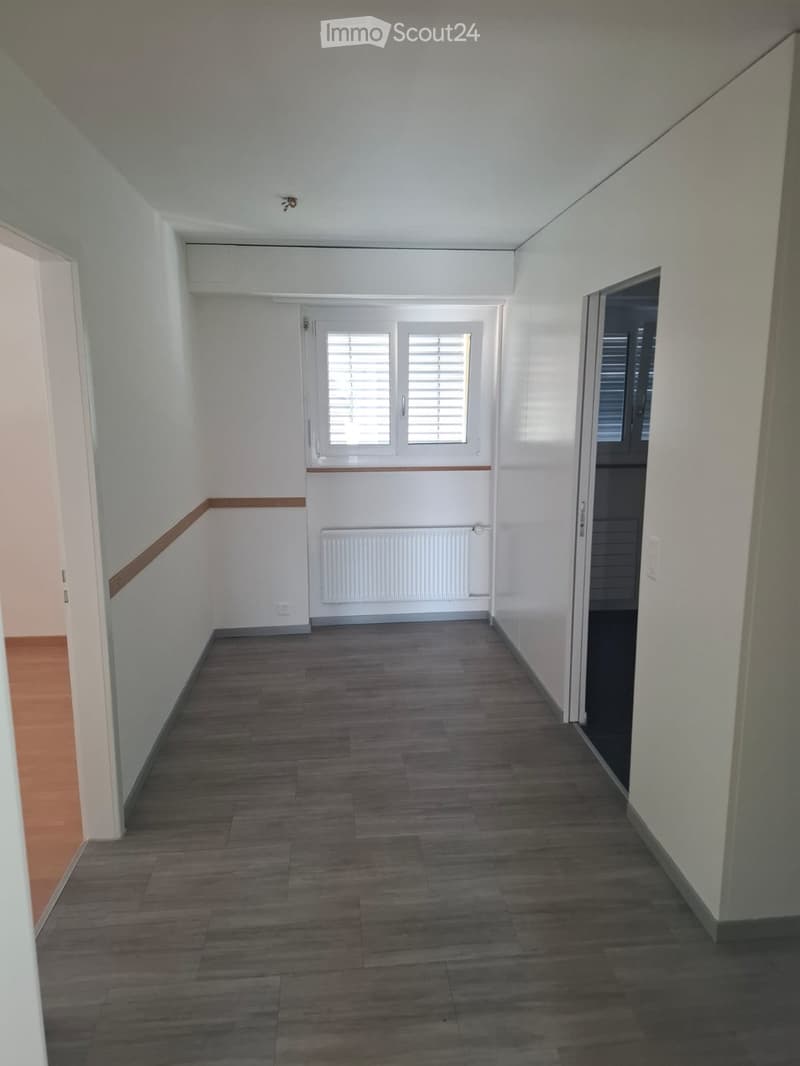 Empty hallway with wooden flooring, white walls, and a radiator. Features a window with blinds and a door on the right side.