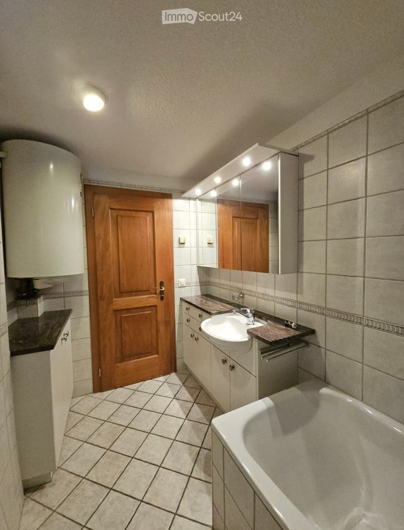 Bathroom with tiled walls and floor, a sink with a mirror cabinet, a bathtub, and a wooden door leading to another room.