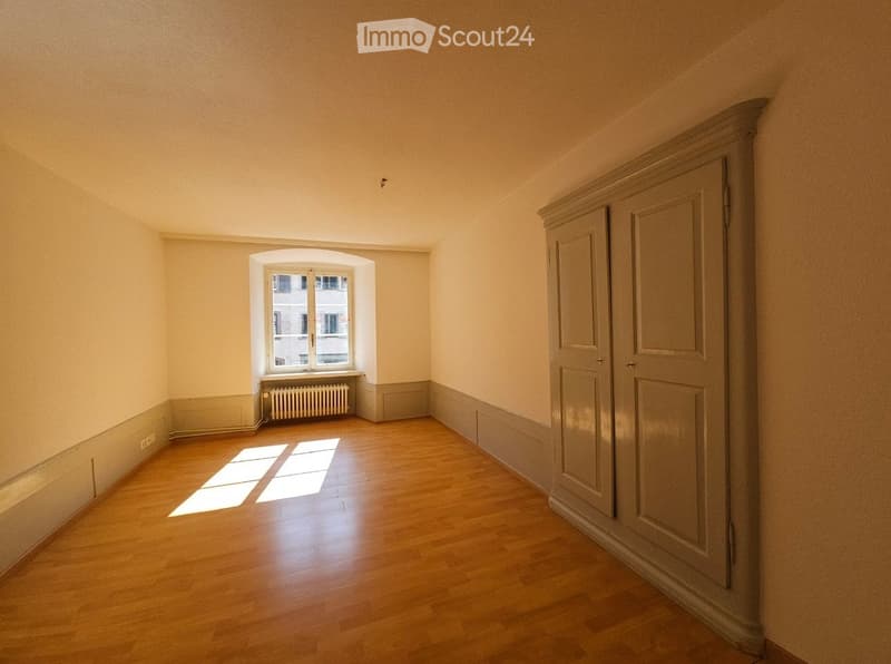 Empty room with wooden flooring, light-colored walls, and a large window with a radiator below it. The room has a built-in cabinet on one side.