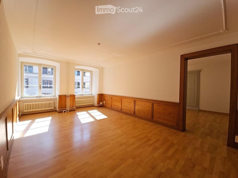 Empty room with wooden flooring, white walls, and large windows allowing natural light. Radiators are visible beneath the windows, and there is a wooden panel wall section.