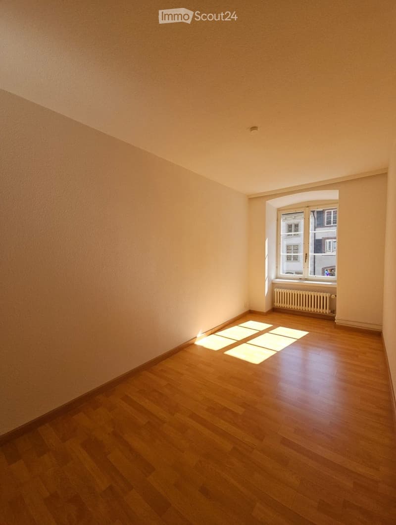 Empty room with wooden flooring, light-colored walls, and a window with a radiator below it.