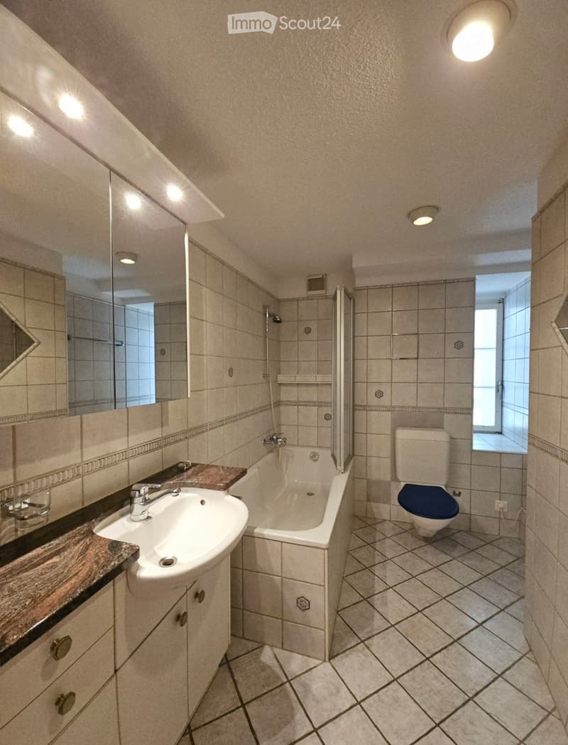 Bathroom with a white sink, bathtub, toilet, and mirror. The walls are tiled, and the floor is tiled with a diamond pattern. There is a window providing natural light.