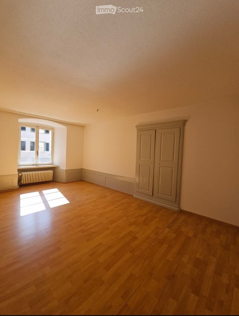 Empty room with wooden flooring, light-colored walls, a window with a radiator below, and a built-in cabinet.