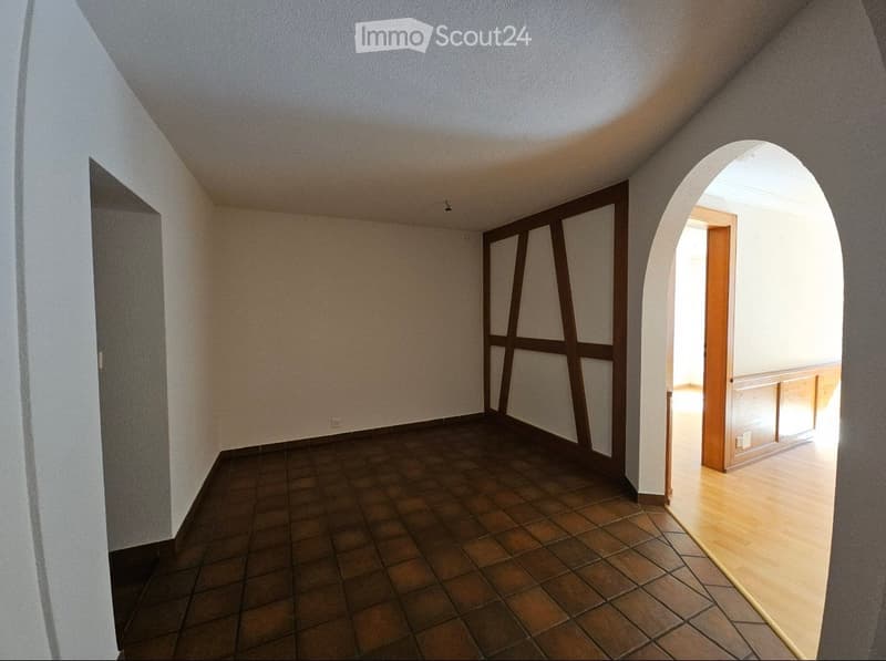 Empty room with beige walls, brown tiled floor, and an arched doorway leading to another room with light-colored flooring.