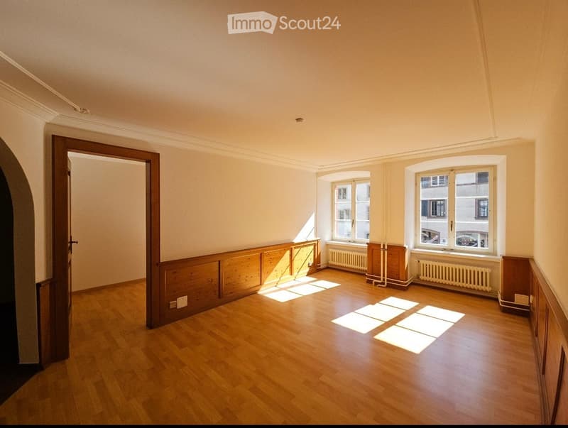 Empty room with wooden flooring, light-colored walls, and two windows allowing natural light to enter. The room features built-in wooden paneling along one wall and a door leading to another space.