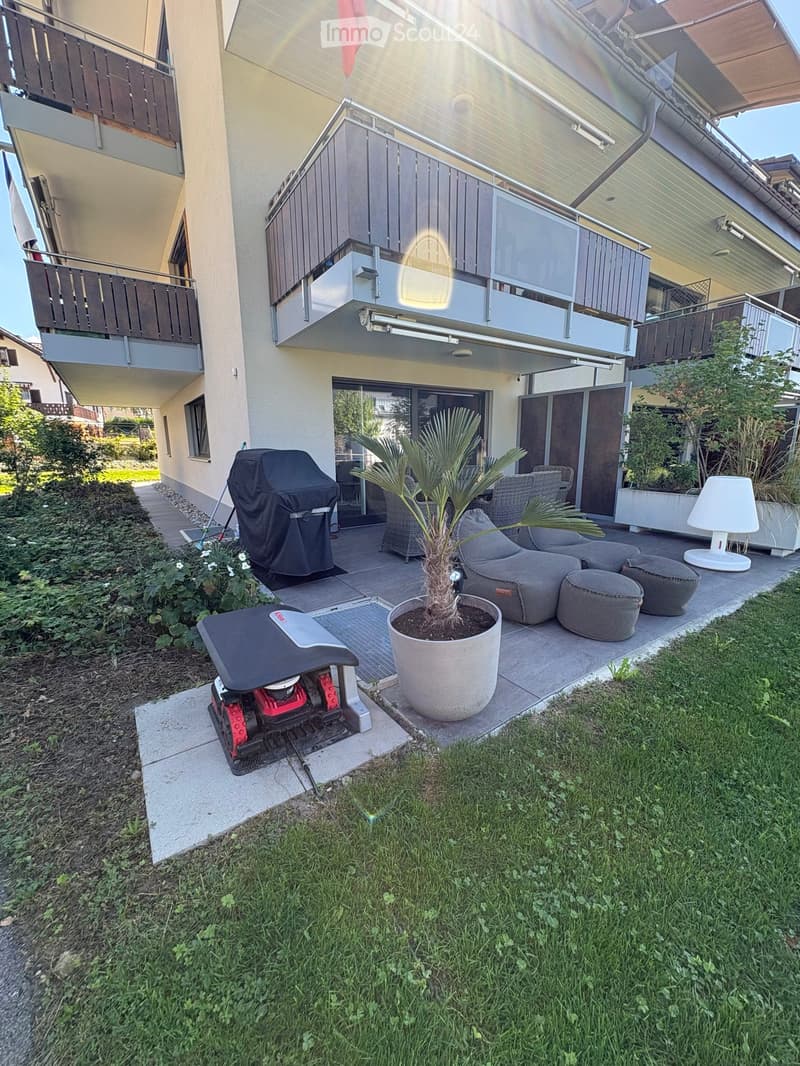Outdoor patio area with a sofa, stools, a lamp, potted plants, and a lawn mower. The patio is adjacent to a modern building with balconies and large windows.