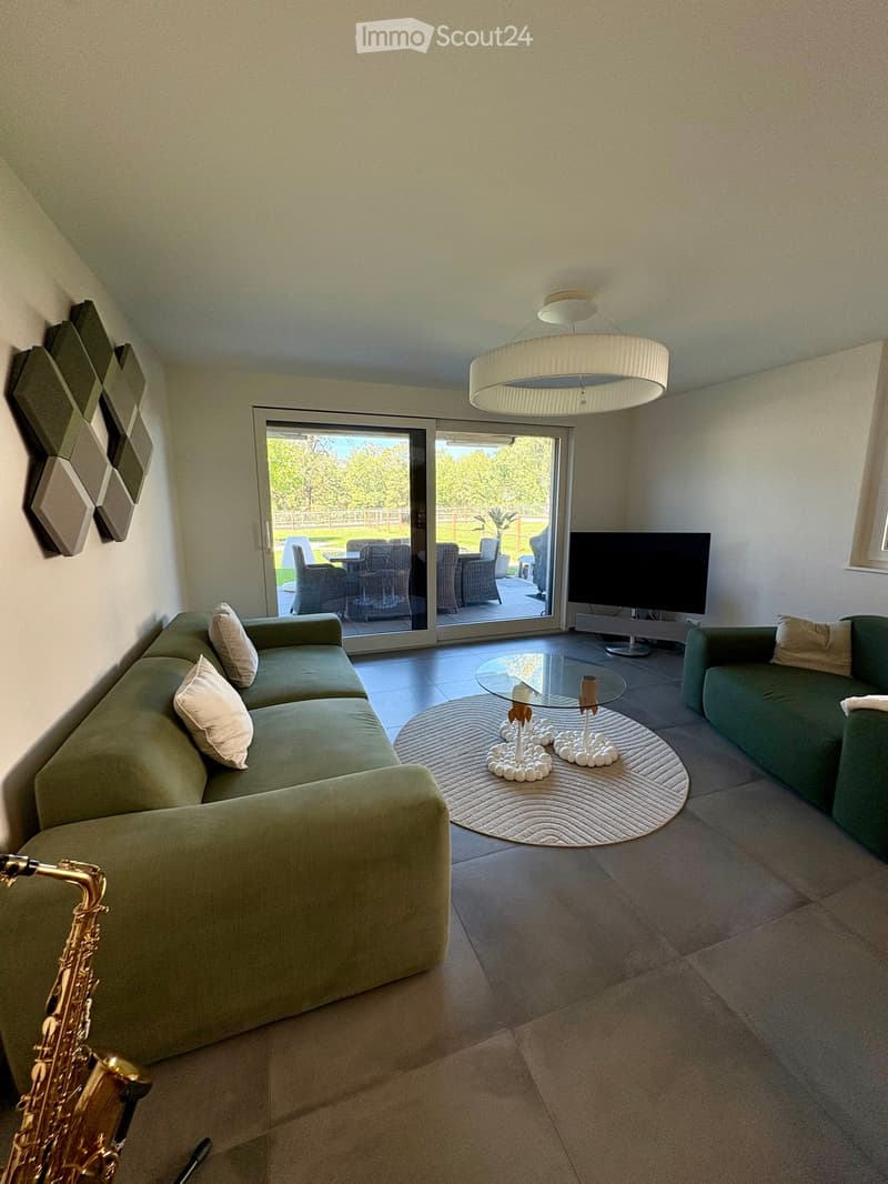 Modern living room with a green sectional sofa, glass coffee table, television, and a view of an outdoor patio through large glass doors.