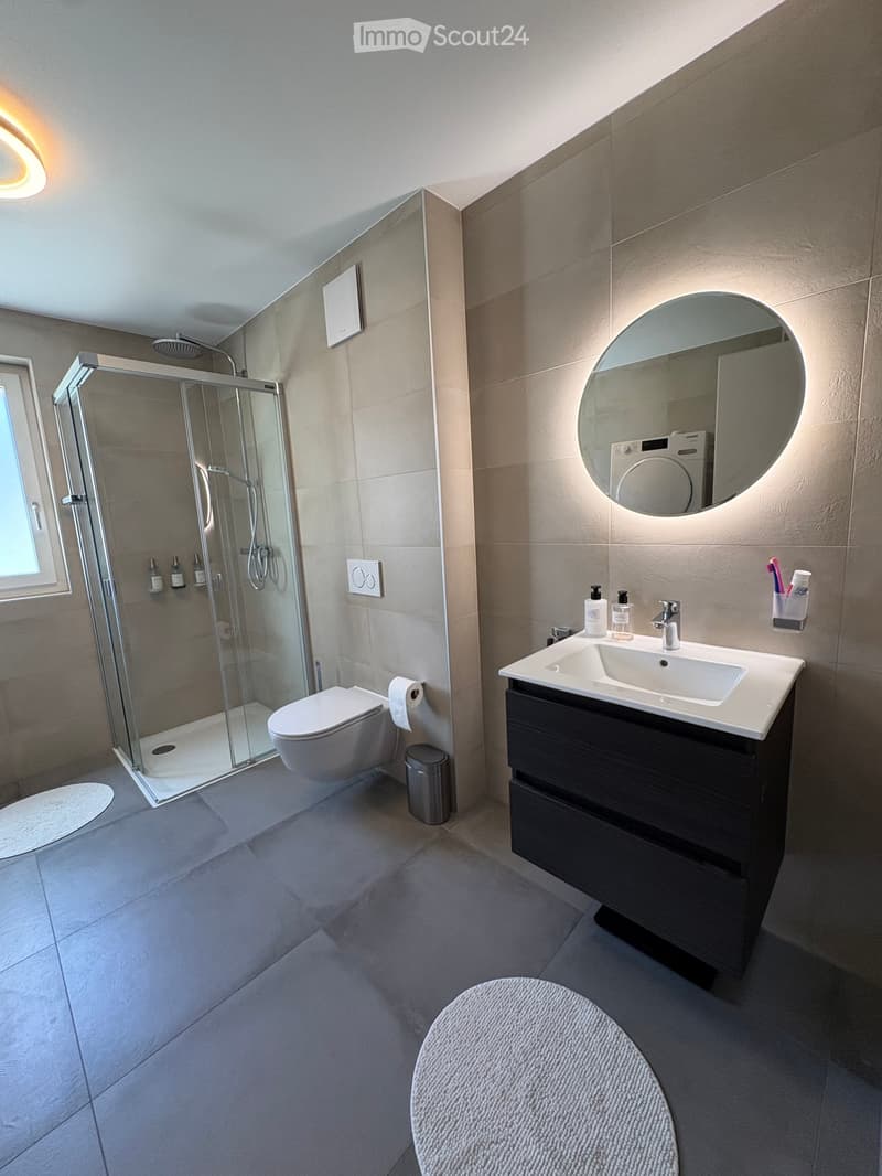 Modern bathroom with a large round mirror, sink with dark wooden cabinet, toilet, glass-enclosed shower, and a small round stool.