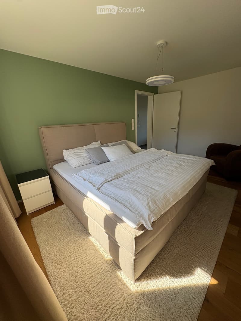Modern bedroom with a large bed, nightstand, and a small chair. The room features a shaggy white rug and a pendant light fixture.
