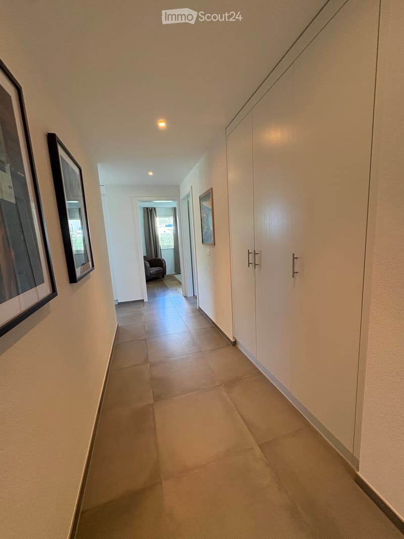 A clean, well-lit hallway with tiled flooring, white walls, built-in wardrobes, and framed artwork. A glimpse of a room with a chair and window is visible at the end.