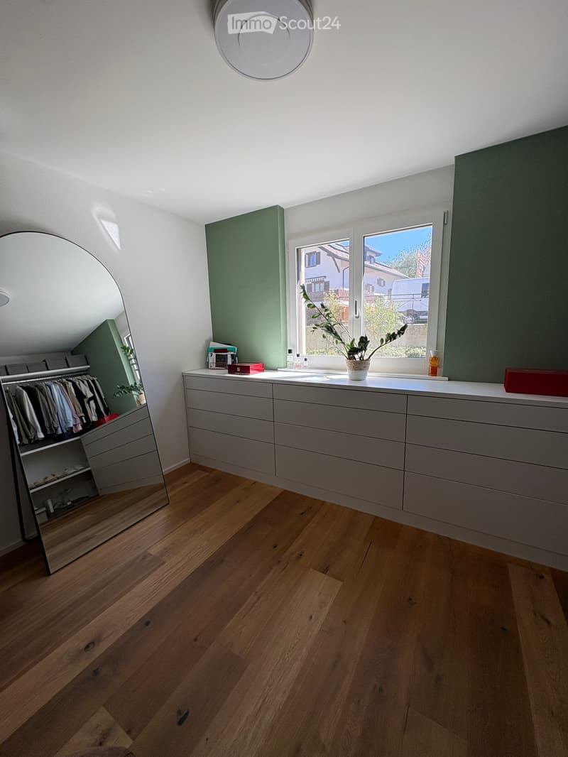 Modern, well-lit room with wooden flooring, a large mirrored wardrobe, a grey drawer unit, and a potted plant on the windowsill.
