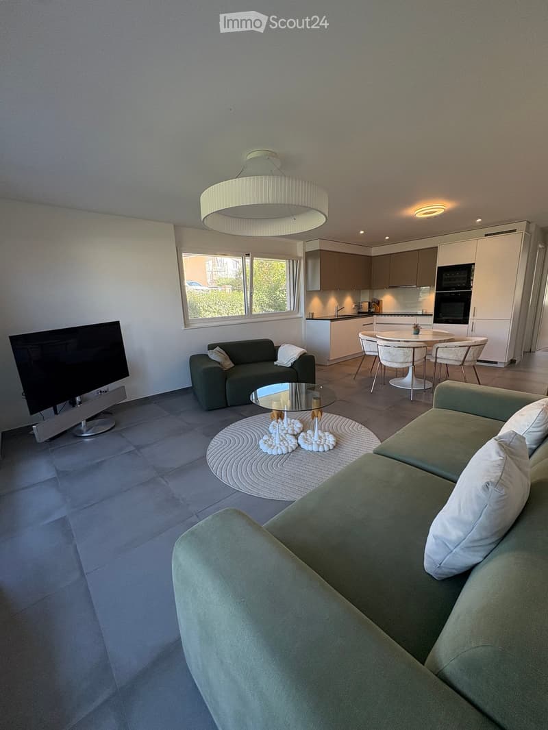 Modern living room with a green sofa, coffee table, TV, and a small kitchen area with a dining table and chairs.