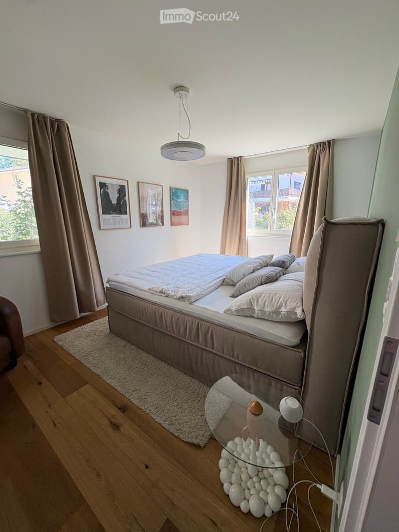 Cozy bedroom with a double bed, nightstand, glass table, and decorative items. The room features wooden flooring, large windows with curtains, and artwork on the walls.