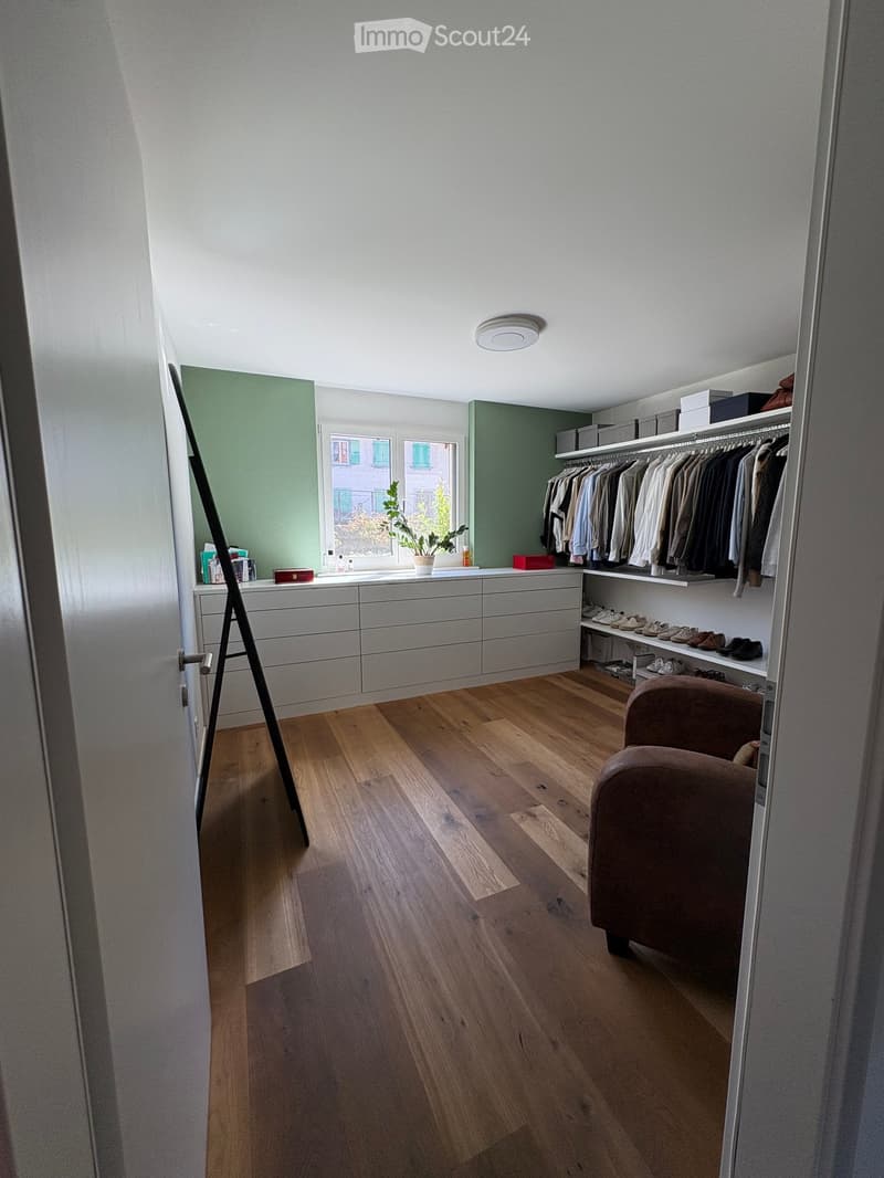 A well-organized walk-in closet with wooden flooring, white walls, and built-in storage including a wardrobe, shoe rack, and drawer unit. A small potted plant sits on the windowsill, and a brown chair is placed near the door.