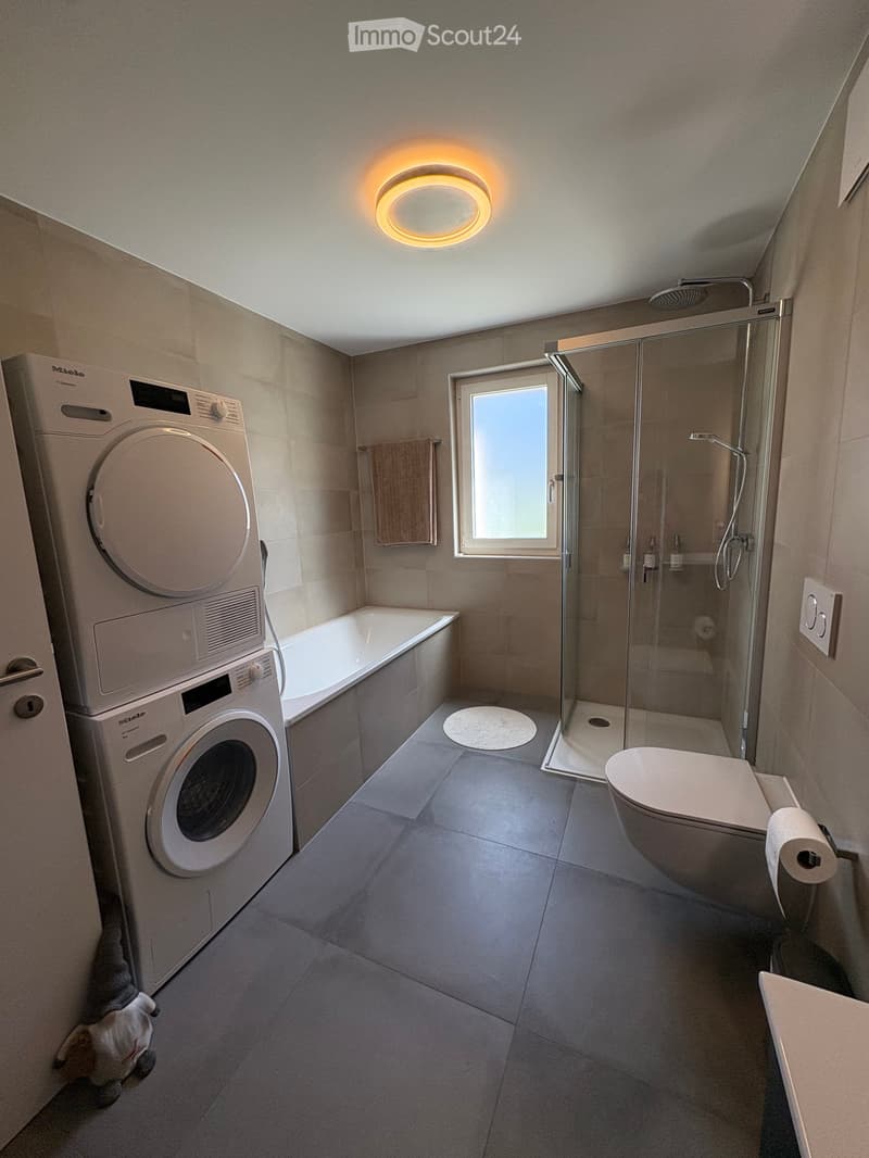 Modern bathroom with a washing machine and dryer unit, bathtub, toilet, glass-enclosed shower, and a towel hanging on the wall.